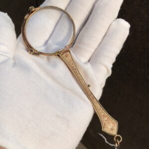 Primer plano de un lorgnette antiguo de oro 14k con detalles esmaltados en el mango, sostenido por una mano enguantada en blanco, resaltando su elegante diseño plegable y ornamentación floral.